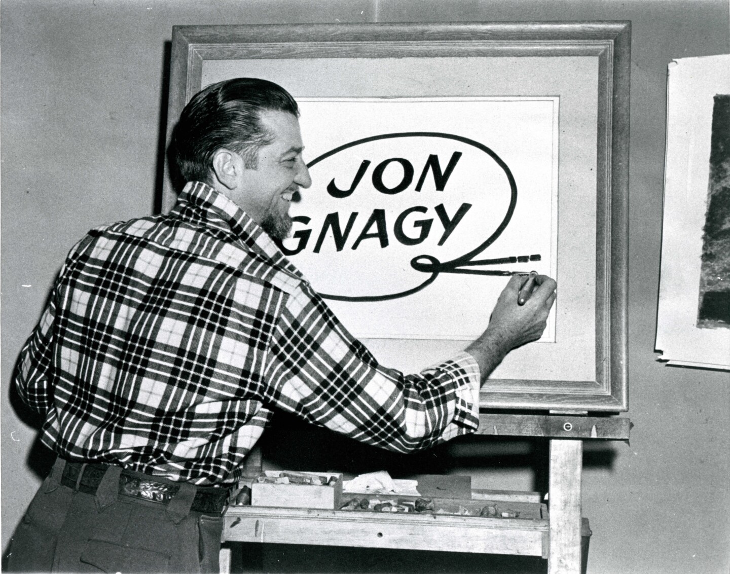 Jon Gnagy signs his name on an easel with his back turned to the camera. The profile of his face can be seen and he is wearing a plaid collared shirt.