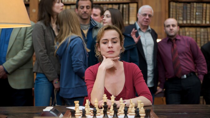 A woman gazes thoughtfully at a chessboard as a crowd of people watch behind her.