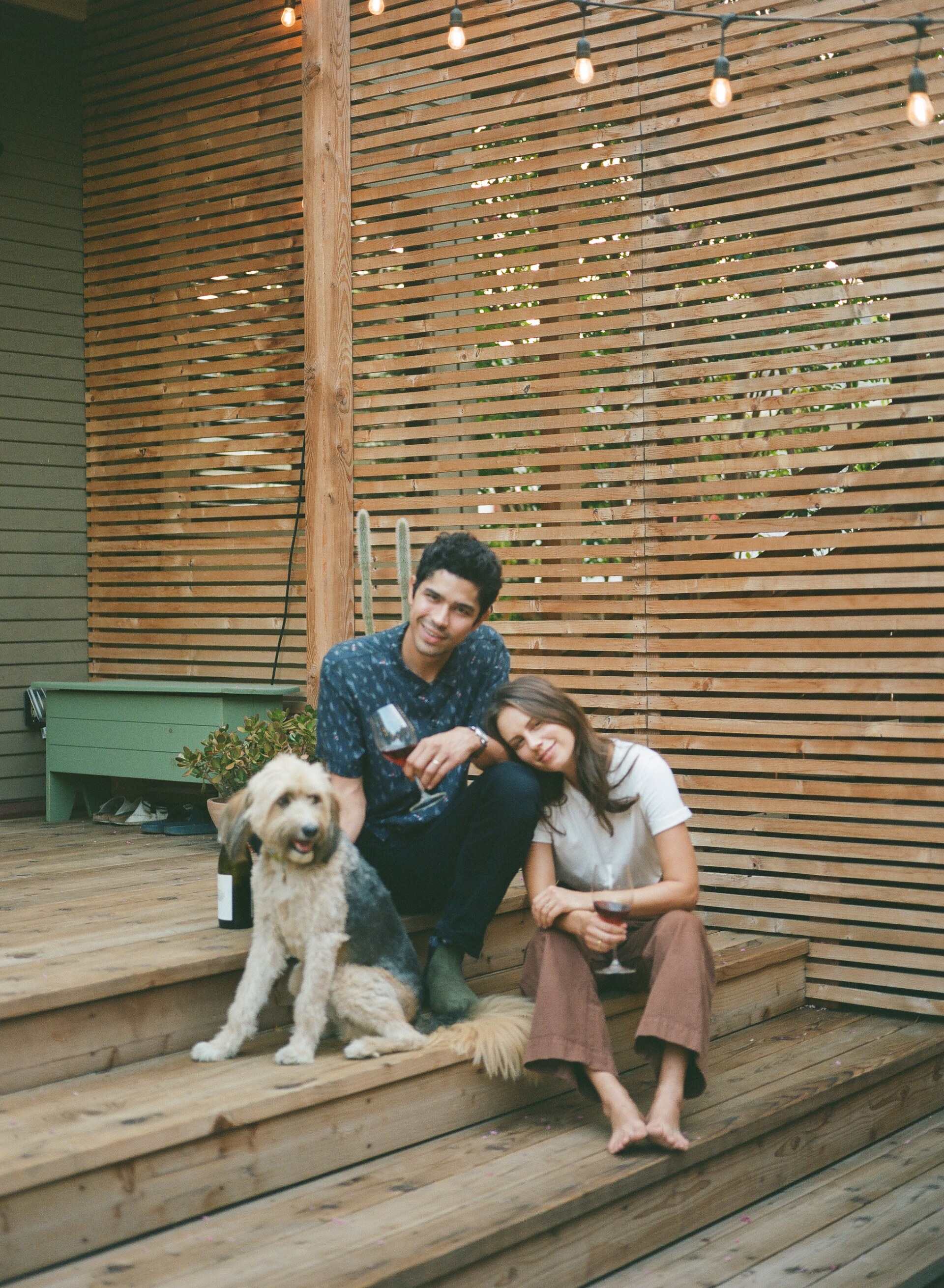 Michel and Rachel DeAscentiis sit on wooden steps, each holding a glass of wine. Next to them is a beige and black fluffy dog. Behind them is a horizontal wood paneling detail.