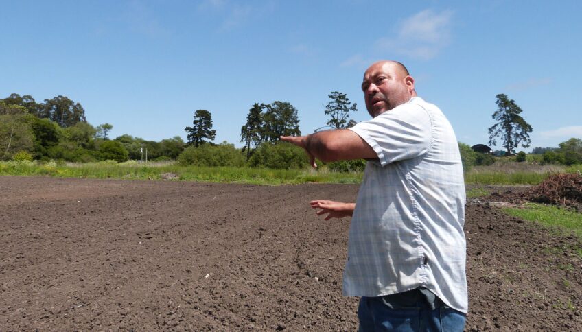 A man with a shaved head and goatee wearing a light-blue buttoned-down shirt points with both arms to a field that had been flooded and now is filled with mud.