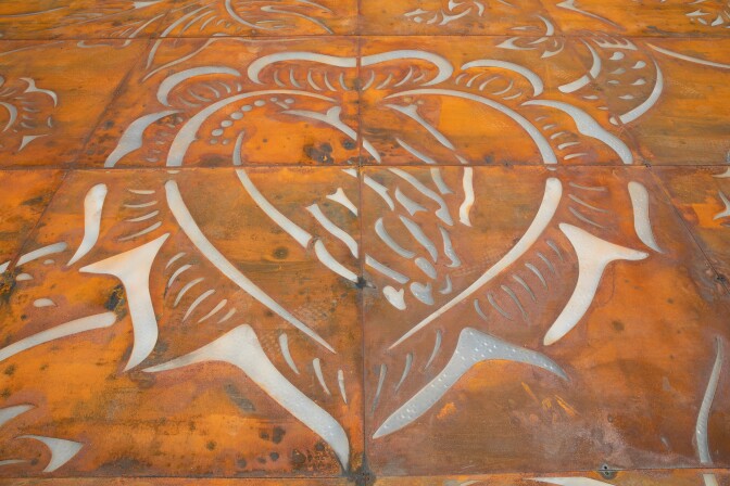 An image of La Virgen de Guadalupe's praying hands framed by a heart is etched onto orange and brown steel plates that resembling weathered leather.
