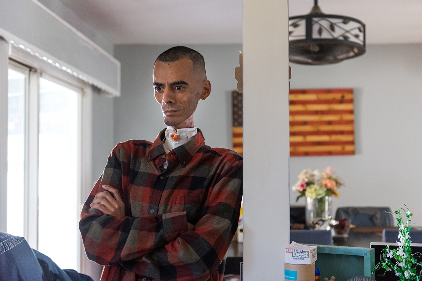 Brian, Rhianna's father, leans against a post in their home. He's standing with his arms crossed, wearing a red button up shirt. Behind him is an American flag wall piece.