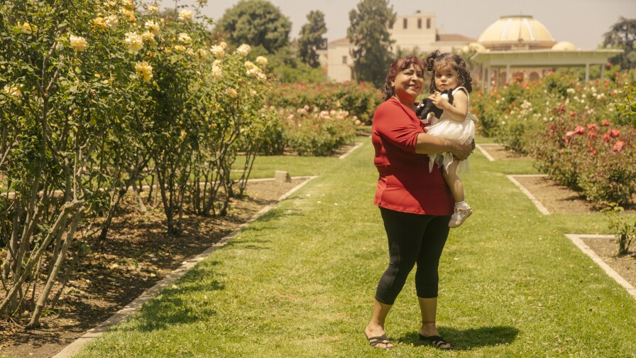 A woman in a red top holds a small girl in a white dress at a rose garden