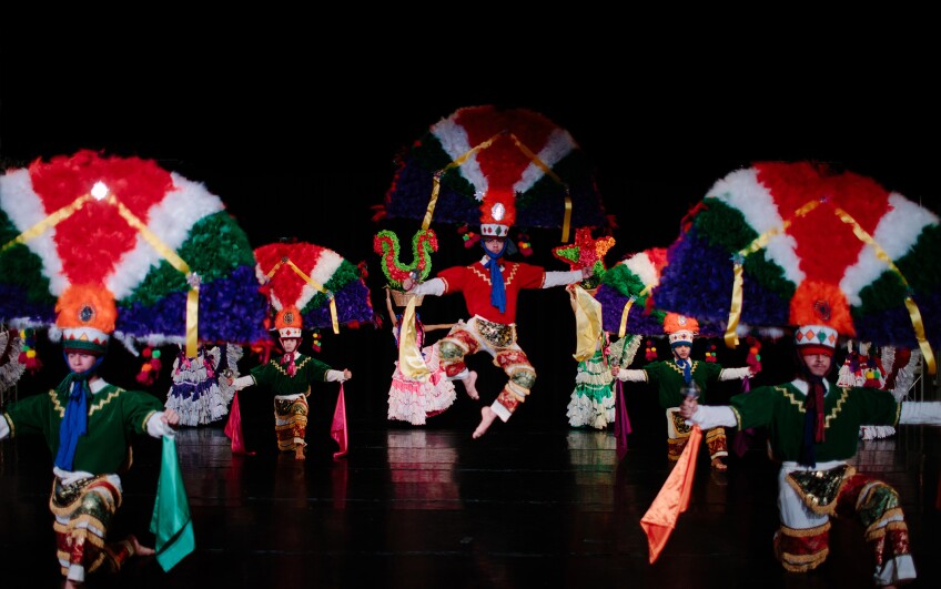 Dance members of Pacifico Dance company are on stage, each wearing large fan-shaped headdresses on their heads. Most of the members are kneeled on the floor in with their arms outstretched to their sides while one member in the center is captured mid-air, jumping with his toes pointed towards one another as he stretches his arms to either side of his body.