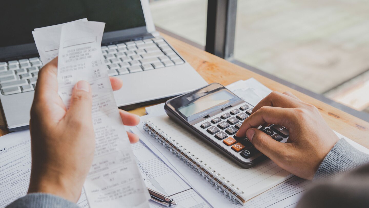 One hand holding a receipt while the other hand punches numbers on a calculator at a desk. | iStock