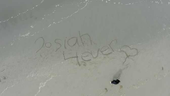 Charmaine Lawson drawing "Josiah 4 Ever" with a heart in the sand on a beach | Still from "Who Killed Josiah?"