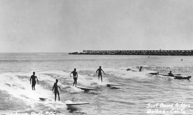 An old, black and white photo of surfers surfing off-shore. A jetty extends behind them. On the bottom left corner, "Hitchcock Photo #55" is handwritten directly on the photo. On the bottom right corner, "Surf board riders Balboa - Calif." is handwritten directly on the photo.