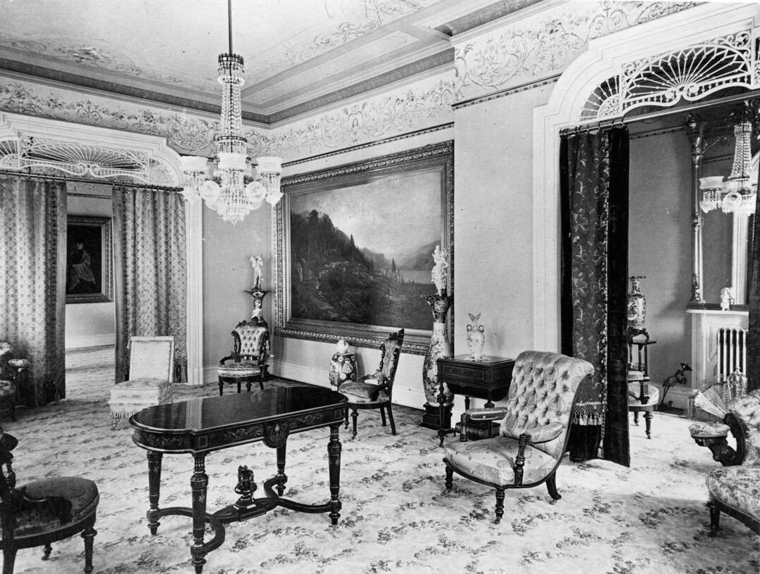 Photograph of a view of the back parlor in the home of Mrs. Arcadia Bandini Stearns de Baker. A small, well-varnished central table is pictured just to the left of the center of the image, surrounded by various plush chairs. A chandelier can be hanging from the ceiling, while a large oil painting of Mount Shasta can be seen in the right background. A painting of Mrs. Baker can be seen through a set of curtains in the background, while to the right, a second, similar room can also be seen through curtains.