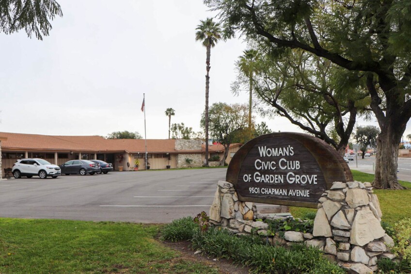 A wooden sign mounted on cobble-stone reads, "Woman's Civic Club of Garden Grove." Below that, "9501 Chapman Avenue." Off in the near distance is a parking lot and a one-story bungalow-style building.  