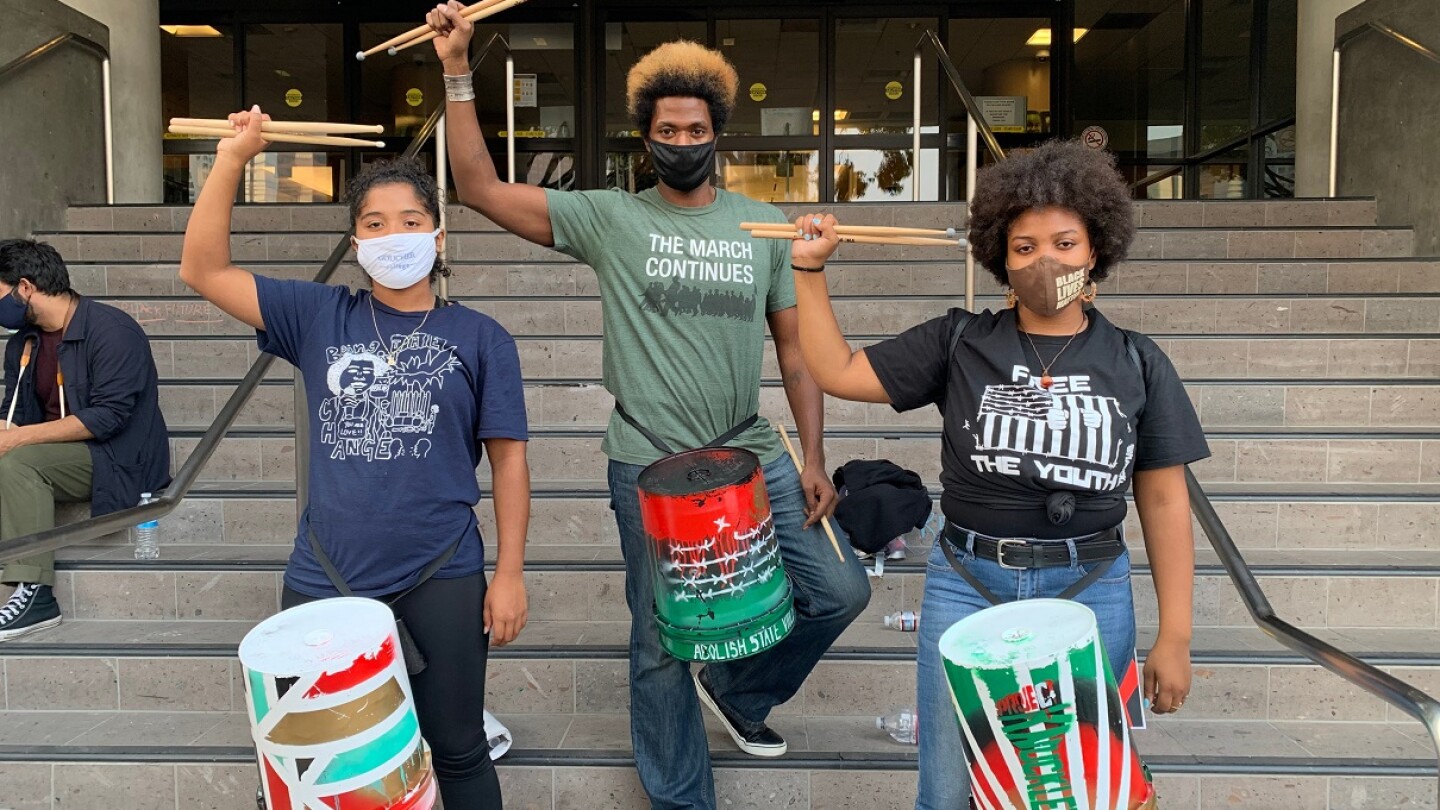 Three young Black Lives Matter protestors hold their drumsticks high in a fist with a drum tied around their waist.
