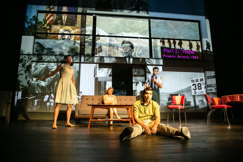 Four cast memebers are on a stage set to look like a living room and dining room. One cast member is sitting on the floor with his legs spread out. He has a sad look on his face. Another cast member is standing in the living room area while another is sitting on the couch with her legs crossed. The fourth cast member stands in between the living and dining rooms. All of the cast members are dressed in midcentury modern clothing, matching the era of the furniture on stage. Behind the stage is a photo collage projected on the wall. Images of John F. Kennedy and war are projected.