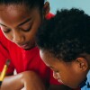Two Black children lean into something they are working on on a table with paintbrushes and crayons.