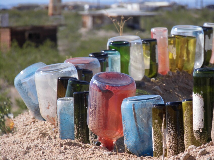 Colored glass bottles of different sizes and shapes are staked into the dirt, side by side, forming a fence of glass bottles. 