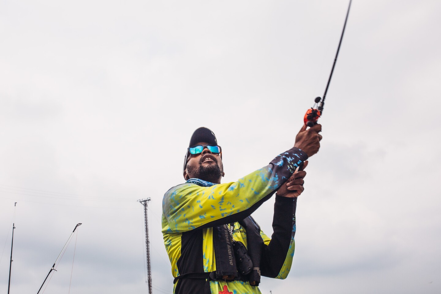 A man casts a fishing line. Behind him is a gloomy, gray-blue sky. The man is wearing sunglasses with teal blue lenses and a bright yellow jacket.
