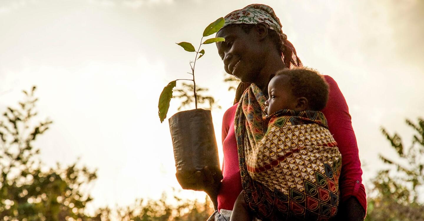 A young mother holding a baby also holds up a young plant in her other hand.