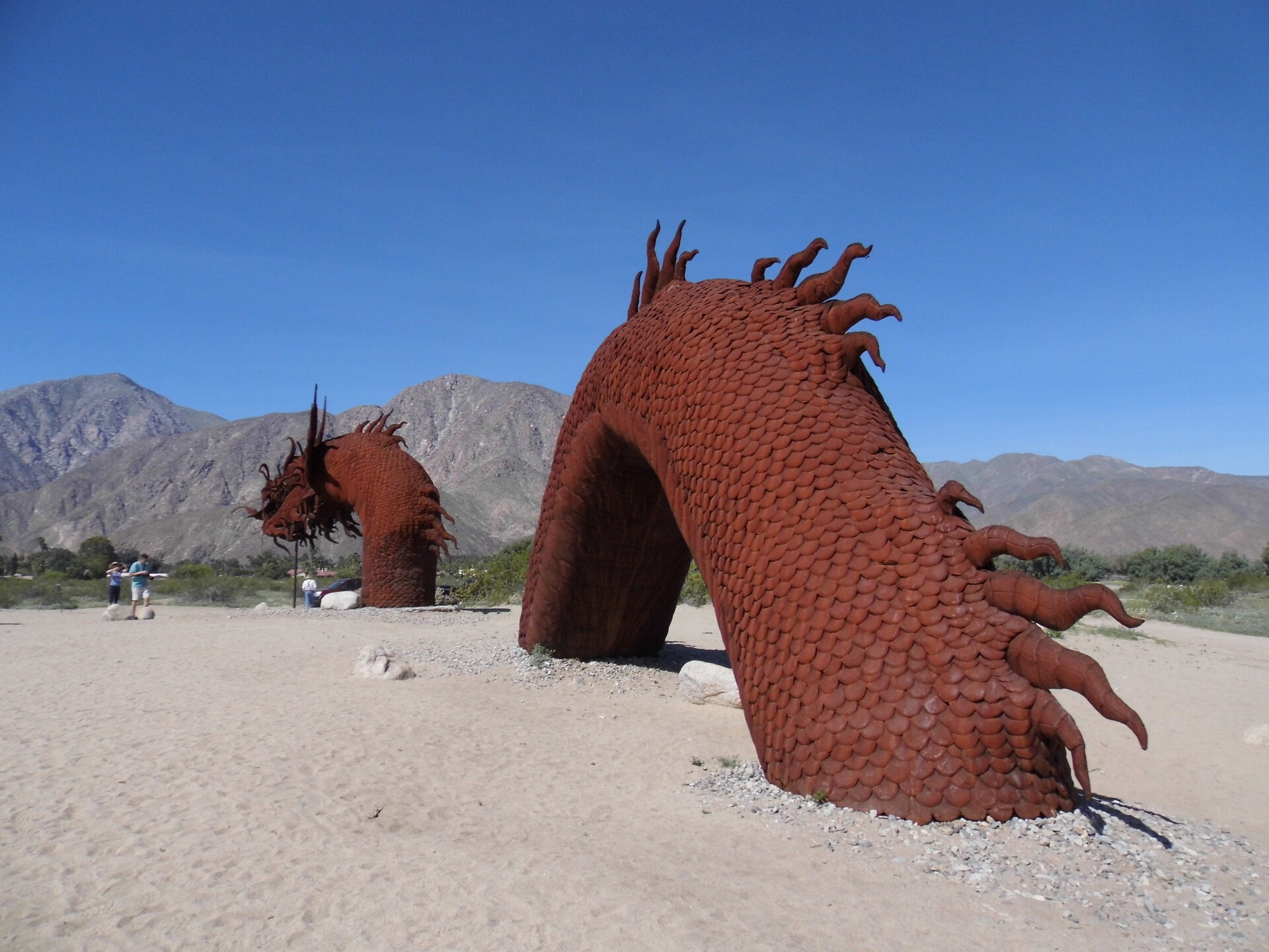 A red iron metal sculpture of a giant serpent emerging from the dirt. In the background, rolling mountain ranges line the horizon. 