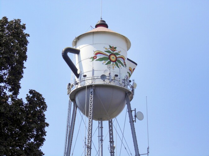 A water tower stands against a bright blue sky. The water tank is shaped to look like a coffee pot, adorned with a handle and a spout. On the face of the water tank is a Swedish floral design.