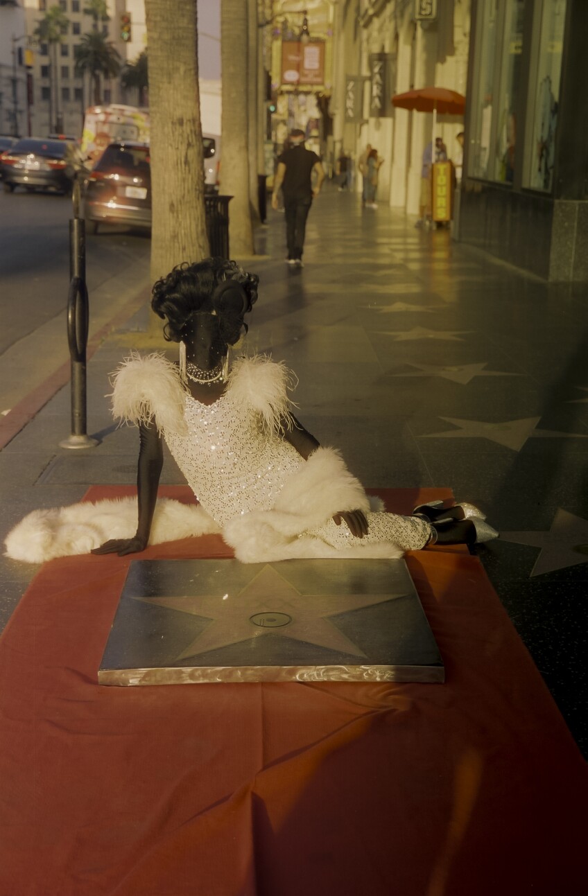 Muxxxe, who is adorned in a black morph suit under a sparkling white dress, curled wig and bedazzled jewelry, sits on a red carpet laid out on the Hollywood Walk of Fame. In front of them is a Hollywood Walk of Fame star recreation sitting atop the red carpet.