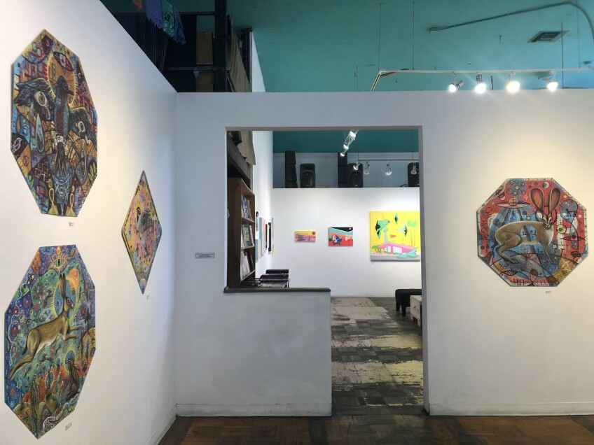 An art gallery with white walls displays wall art in the shape of octagons and diamonds. An upside-down L-shaped doorway reveals another part of the gallery where there is a bookshelf and other wall art. 