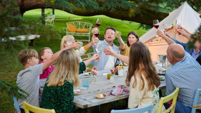 Jamie Oliver toasts his glass with a group of people at a dinner table.