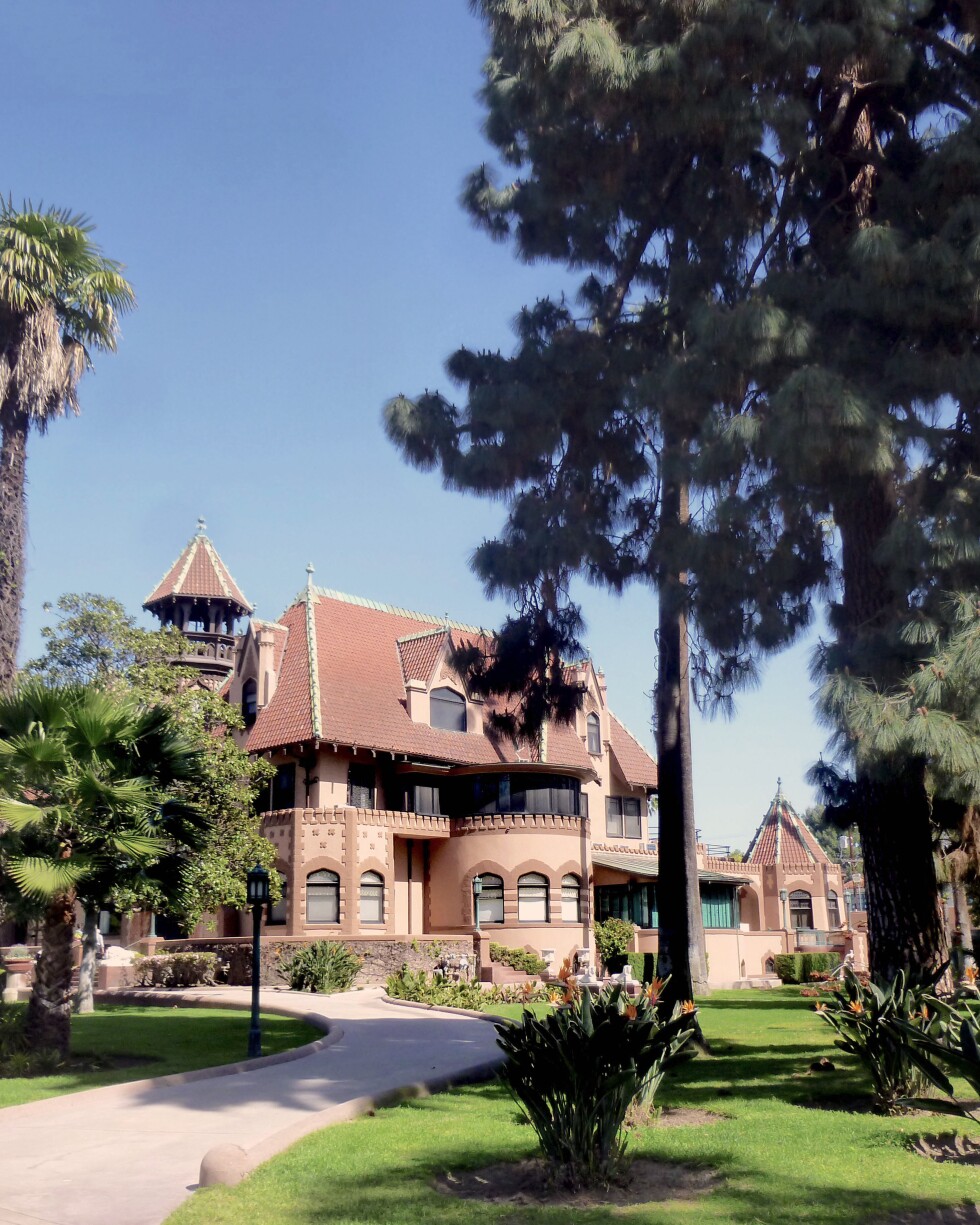 The Doheny mansion, an red-orange brick building of French Chateauesque style architecture, mixed with Gothic, Moorish and California Mission elements stands among a grove of various trees. A paved pathway winds up to the mansion. Above the mansion is a bright blue sky.