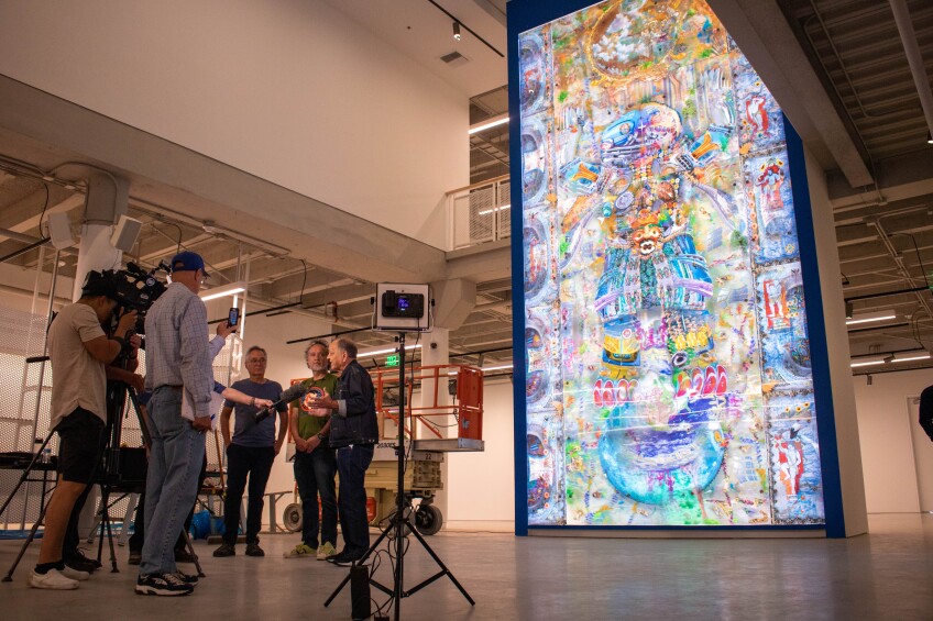 Three men being interviewed in front of a large LED-backlit artwork.