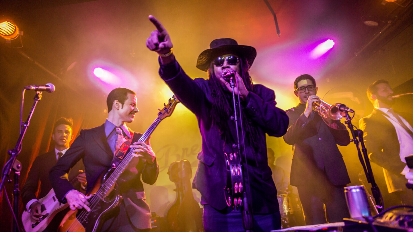 A band, composed of (from left to right) a guitarist, a bassist, a singer, a trumpet player and a saxophone player, performs on a stage. They're all wearing suit jackets and playing their respective instruments. The singer in the middle is pointing out to a crowd. Behind them are neon pink and yellow lights that provide a hazy glow in the backdrop.
