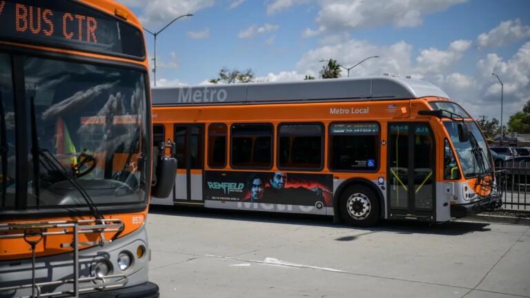 The front of an orange and gray city bus is in the foreground with a wider shot of another Metro Local orange and gray city bus pulling up alongside it in the background
