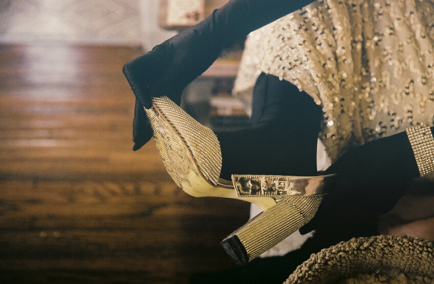 Muxxxe, who is wearing a black morph suit under a sparkly, glamorous dress puts on a silver bedazzled high heel.