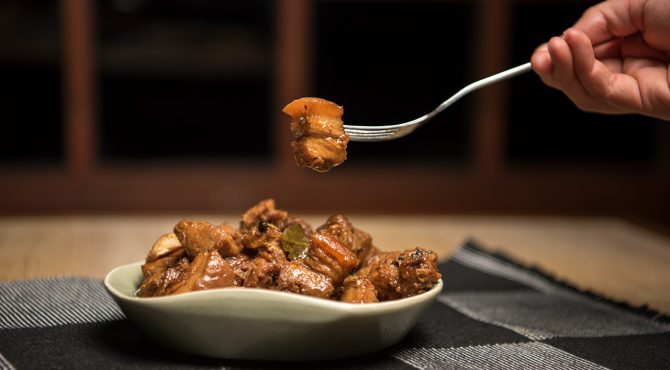 A hand raises a piece of meat from an adobo dish with a fork.