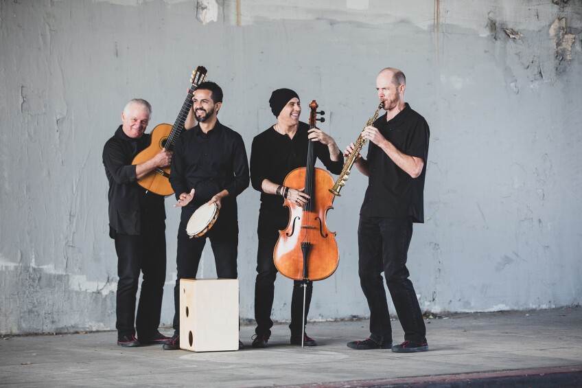 Four men stand in front of a distressed gray wall. The man on the left is holding up an acoustic guitar, next to him a man is holding a tambourine while a light wood-grain cajón sits at his feet, next to him a man wearing a black beanie is holding a cello and next to hiim a man is playing a brass soprano saxophone.