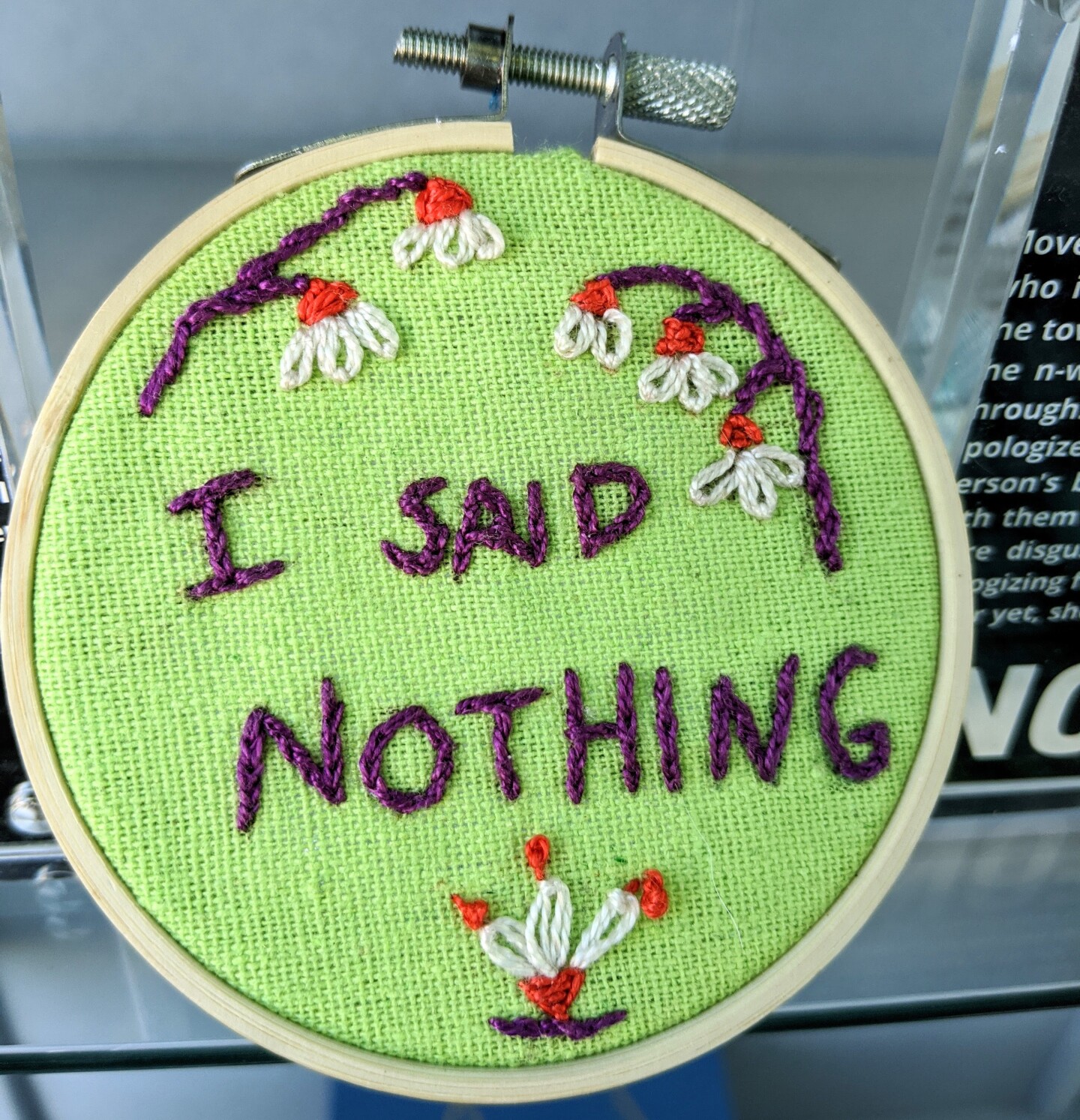 A small embroidery hoop with bright green fabric stretched across embroidered with the words, "I said nothing" in purple thread. Surrounding it are floral designs featuring white petals, orange details and purple branches.