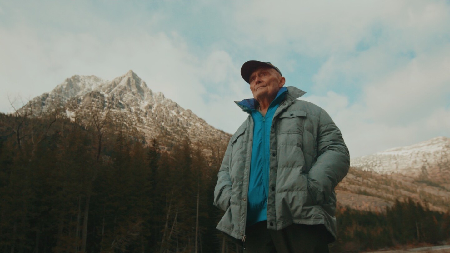 An elderly man stands wearing a powder blue puffer jacket and a teal windbreaker underneath. His hands are in his pockets and a baseball cap sits on top of his head. Behind him is a gorgeous landscape consisting of a towering, snowcapped mountain to his left and a forest of trees of various shades of greens, orange and yellow.