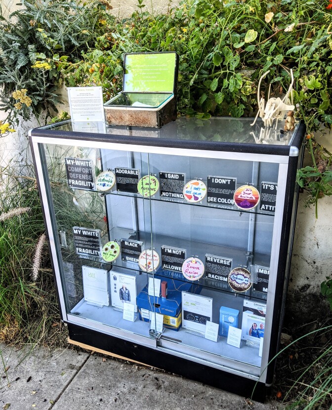 Eight embroidery designs of various colors and phrases centered around the theme of white fragility sit behind a three-shelf glass display. Behind it is a concrete wall covered in thick green foliage.