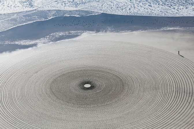 An aerial shot captures massive circles drawn into sand, radiating from a singular point in the landscape. For scale, a long shoreline extends beyond the artwork and a man is seen at the corner of the frame — a mere speck in relationship to the artwork.