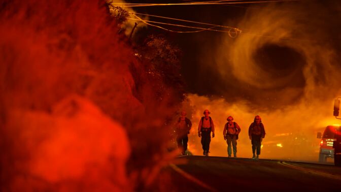 Four firefighters walk down a road with smoke spiraling in the background.