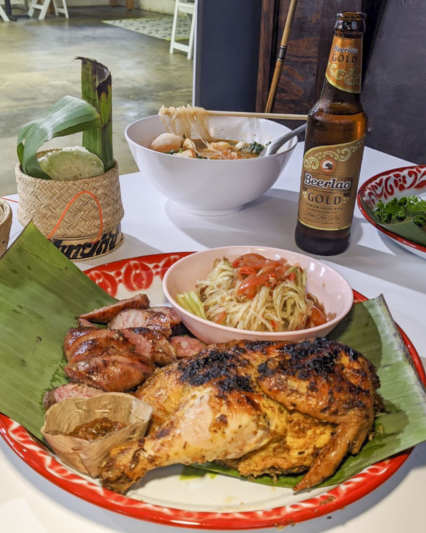 A table is prepared with various Lao dishes. The plate in the foreground is a papaya salad with Lao sausage and chicken. Behind it is a bowl of sukiyaki. To the left of the papaya salad is sticky rice nestled in a small woven basket and to the right is a plate of sakoo yut sai or tapioca dumplings. A bottle of beer also sits on the table.