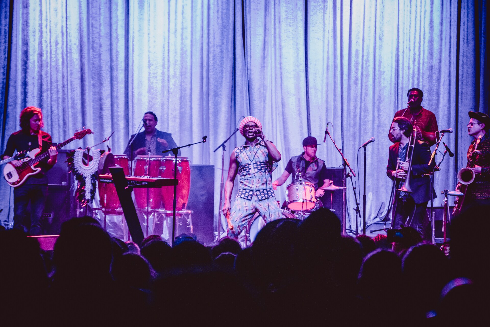 A band performs on a stage lit by a neon blueish-purple light overhead. A singer stands in the middle holding a microphone. They are wearing a vibrantly colored and patterned vest and pants set and orange headwear. Behind the lead singer are various members of the band — (from left to right) a guitarist, percussionist, drummer, trombonist, backup singer and saxophonist. At the foreground of the image is a silhouette of a densely packed crowd watching.