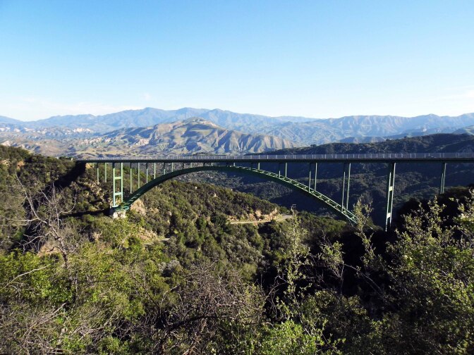 A green arch bridge made of metal beams stretches between two mountains. Behind it are rolling mountains and a clear blue sky.