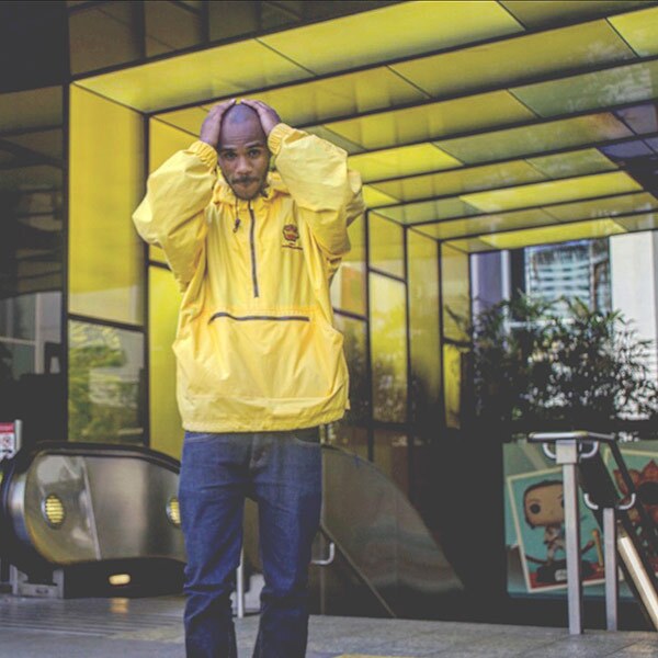A young person wearing a yellow windbreaker holds their head in their hands while posing in front of a yellow glass structure.