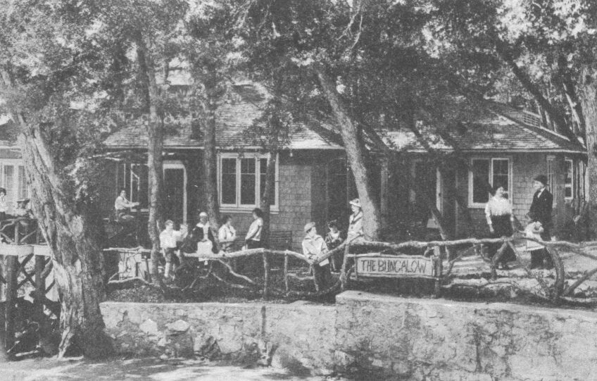 A black and white archival photo of the "Bungalow" at Mt. Lowe. Trees shroud a tiled one-story building. Patrons sit and lounge around an outdoor patio area. 