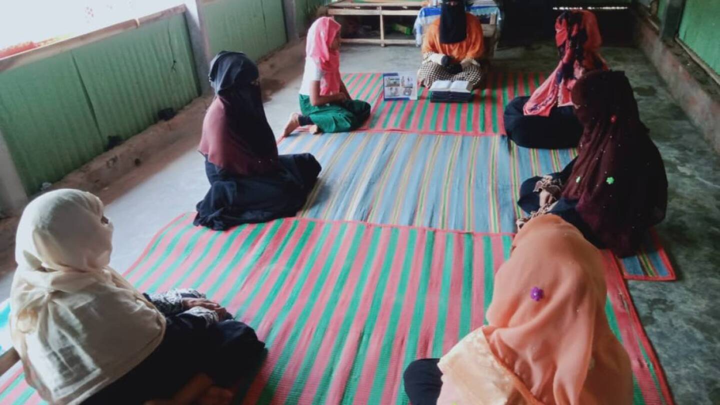 A woman in a burka sits down on the floor as she speaks to several other woman who are dressed and seated in the same way.