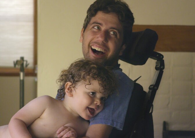 Ady Barkan, an activist living with amyotrophic lateral sclerosis (ALS) and the subject of the documentary 'Not Going Quietly,' smiles while holding his son on his lap.