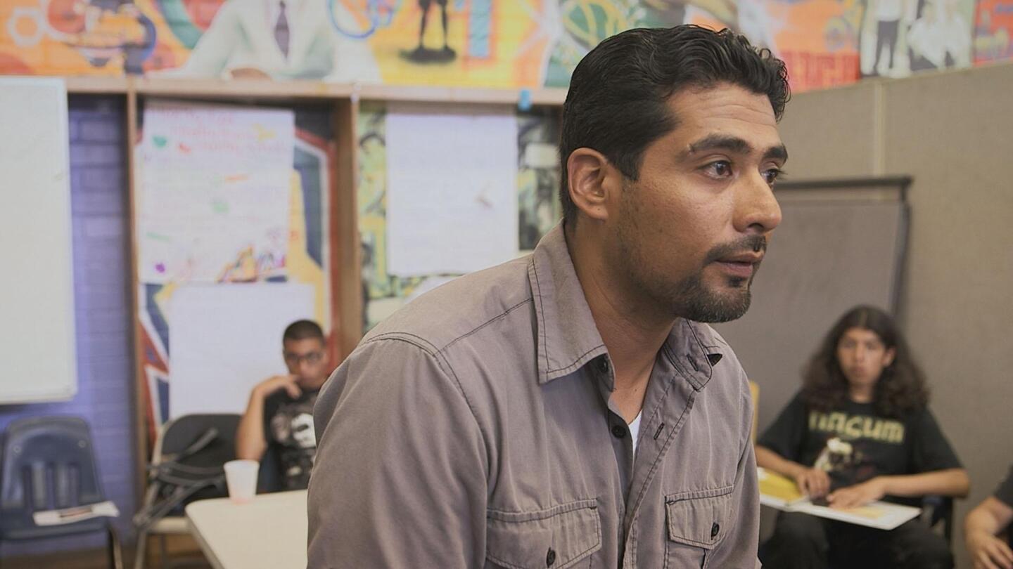 A man in the middle of a room speaks to children sitting in desks. | From "VOCES: The Pushouts"