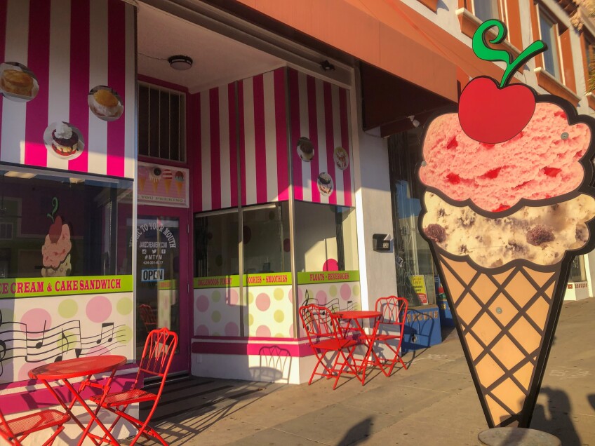 An ice cream storefront with bright red tables and chairs on the sidewalk. A large sign shaped like two scoops of ice cream on a waffle cone and topped with a cherry stands by the door. The store itself is brightly colored, with pink and white stripes and images of ice cream all over. 