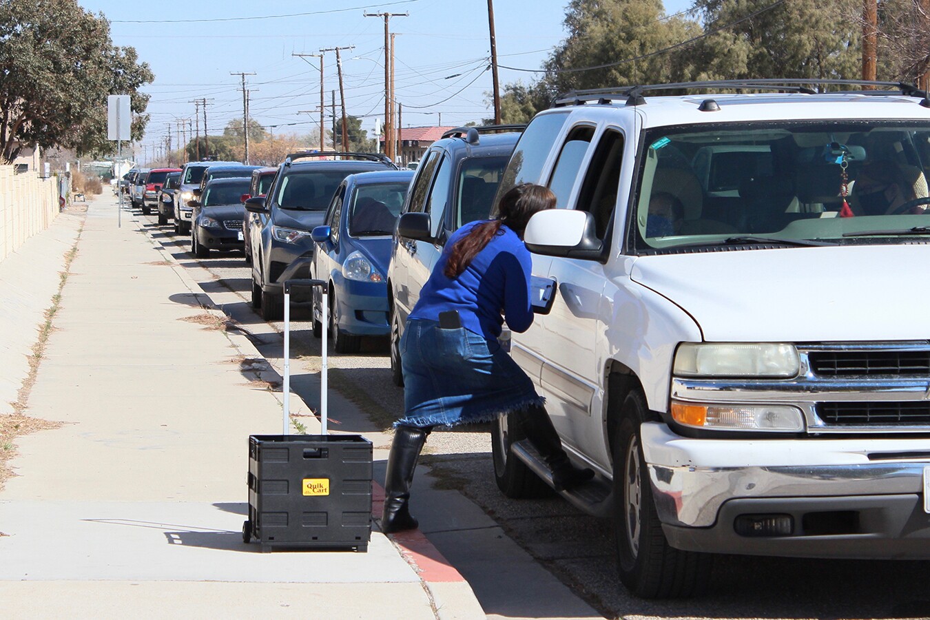 A woman leans into the window of a white SUV at the front of a long line of vehicles.