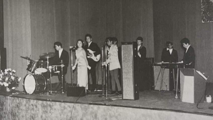 A black and white photo of Thee Chozen Few performing on a stage. There are two people standing in front of microphone stands while others surround them with a drum set, electric guitar, bass guitar and keyboard. Tall speakers are dispersed throughout the stage.