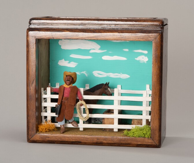 A small shadowbox miniature diorama with wooden frames depicts a scene of a Black cowboy and Oklahoma rodeo champion Bill Pickett leaned against a white fence. He has a lasso in one hand and a cowboy hat atop his head. Behind the fence he's leaning on is a horse.