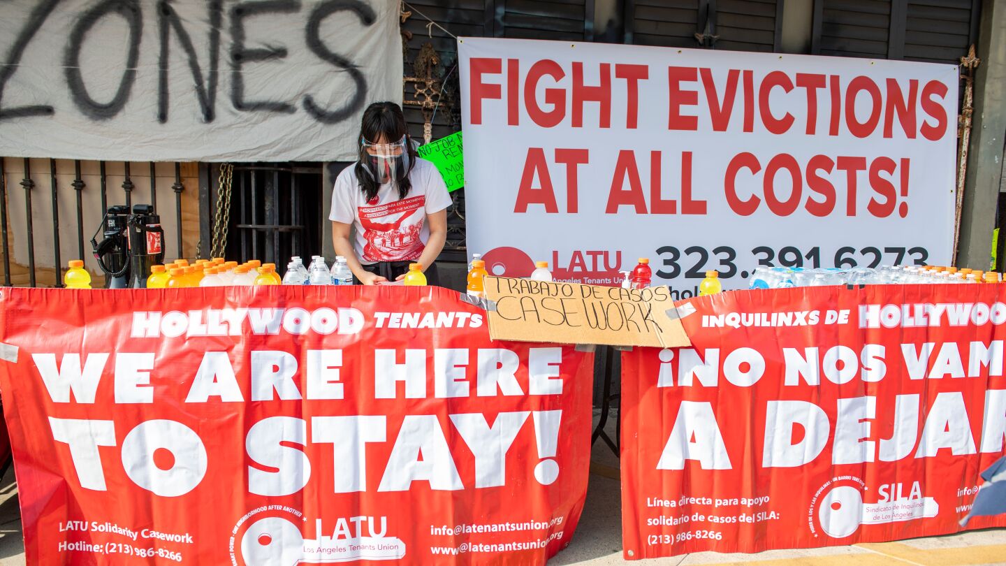 A woman wearing a mask standing behind a table covered in large banners that say "We are here to stay!" and "Fight evictions at all costs!"