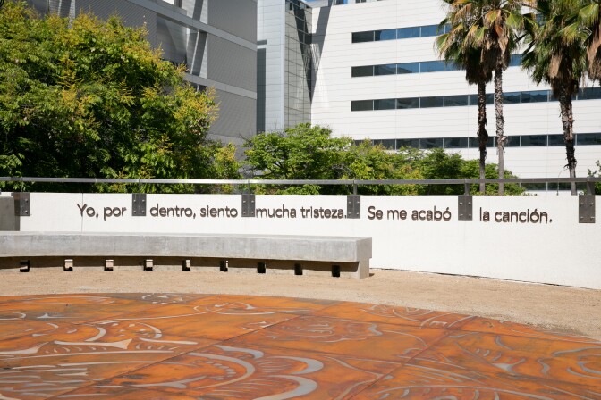 Benches are placed against a half wall where the words, "Yo, por dentro, siento mucha tristeza. Se me acabó la canción," are mounted onto. In front of the benches are metal tiles forming a larger artwork on the ground. Etchings of flowers and folded hands are all over the art piece.